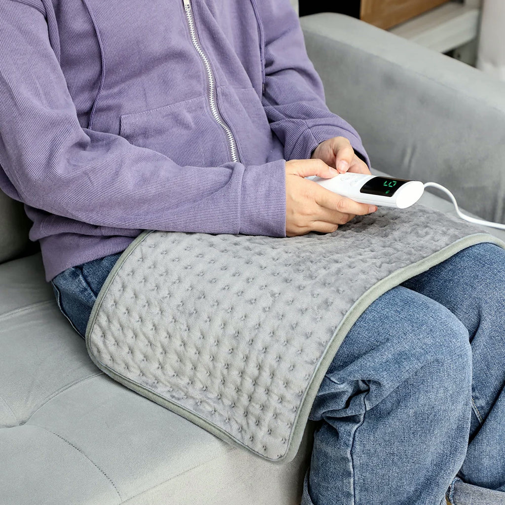 Coussin chauffant gris posé sur les genoux, télécommande à écran en main, assise sur un canapé