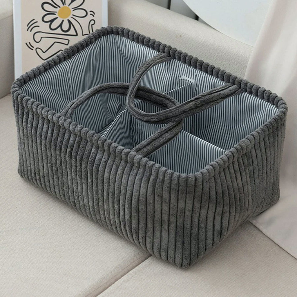 Panier de rangement en velours côtelé gris foncé posé sur un canapé, intérieur rayé à compartiments et anses