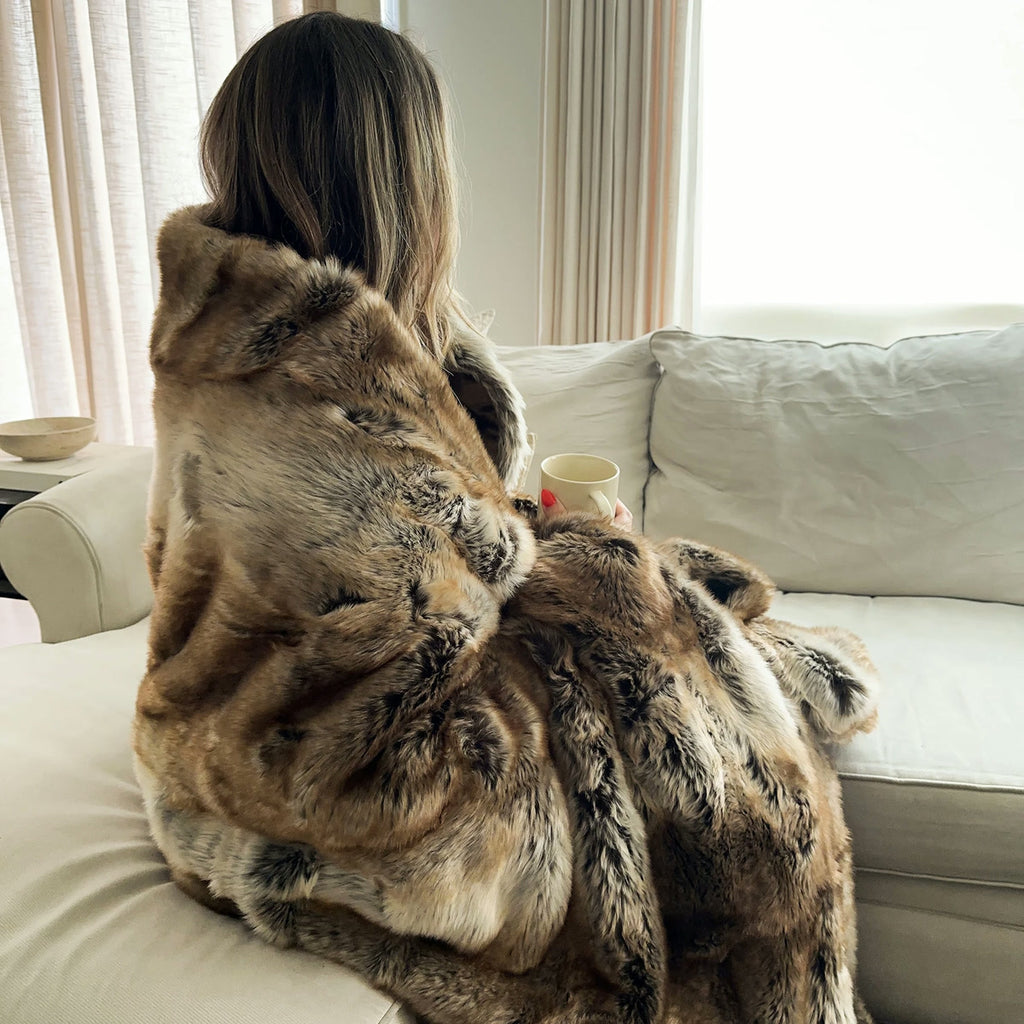 Femme de dos sur un canapé blanc, enveloppée dans le plaid fausse fourrure, une tasse à la main