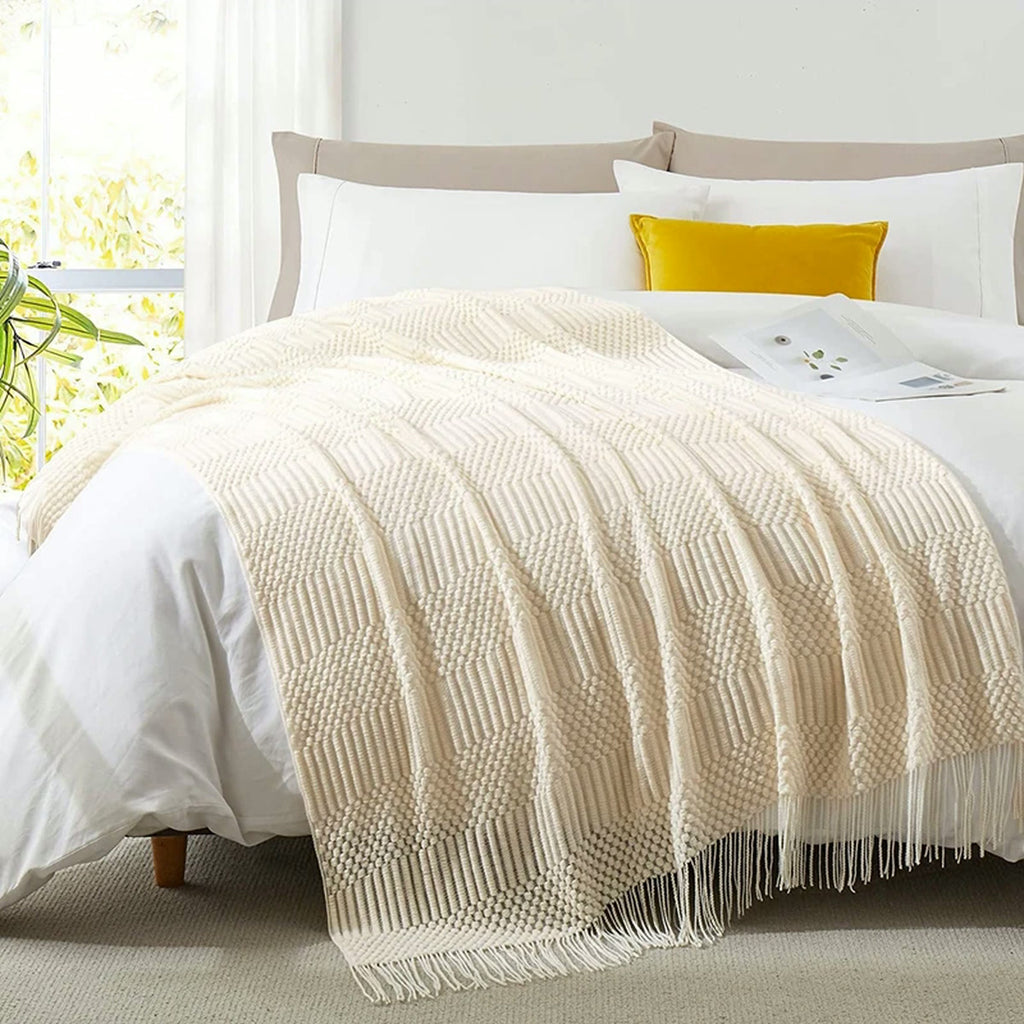 Plaid tricoté écru posé en travers d'un lit blanc, franges tombant au pied du lit