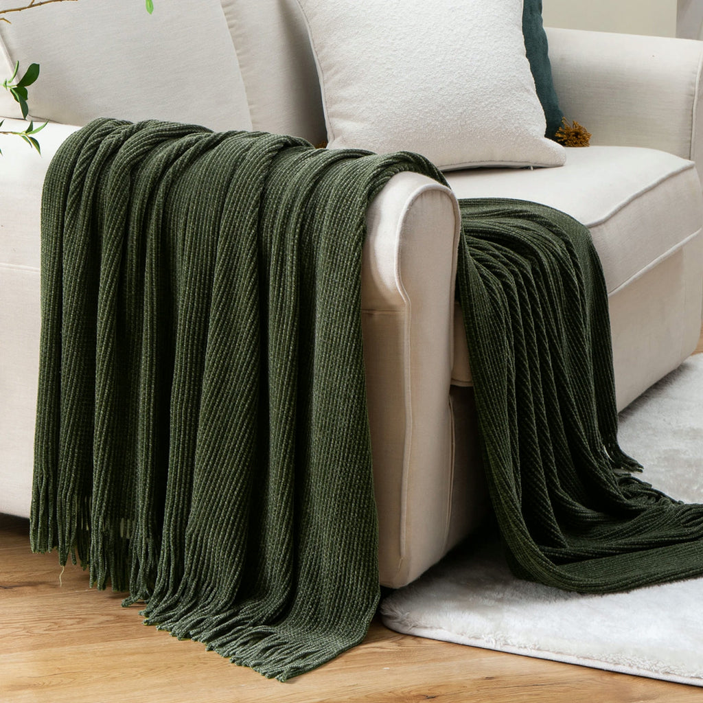 Plaid tricoté vert sapin drapé sur un canapé crème, franges tombant sur un tapis blanc